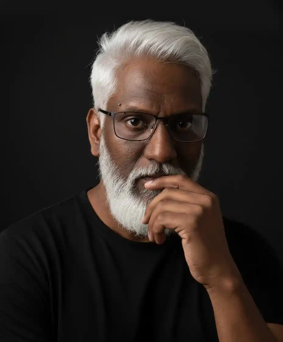 Fernando Kylas, photographed against a black ground, one hand raised to his chin, looking directly at the camera.